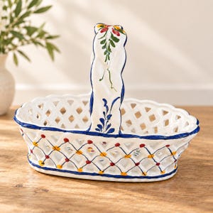 May include: A white ceramic basket with a lattice-style openwork design, featuring a tall, arched handle. The basket is decorated with hand-painted blue trim along the edges and small red and yellow floral accents at the lattice intersections.