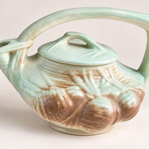 May include: A ceramic teapot featuring a light green glaze with brown accents. The body is molded with a relief pattern of root vegetables. It has a single arched handle extending from the spout to the back and a matching removable lid.