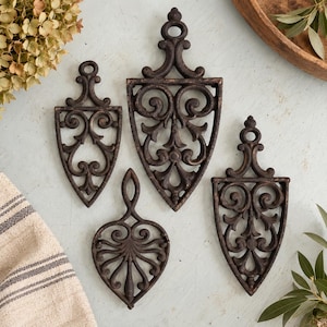 May include: A set of four dark brown cast iron trivets featuring intricate scrollwork and openwork designs. Three of the trivets have a pointed, shield-like shape with a top handle, while one is rounded with a central stem and symmetrical curves.