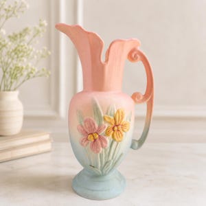 May include: A ceramic pitcher vase featuring a pastel gradient of peach, pink, and light blue. The body is decorated with raised, three-dimensional floral motifs in pink and yellow with green leaves. It has a curved handle and a scalloped rim.