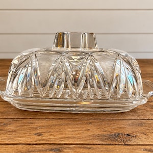 May include: A clear glass butter dish featuring a rectangular base with side handles and a removable domed lid. The lid is decorated with a geometric pressed pattern of vertical rays and cross-hatched textures, topped with a rectangular knob.