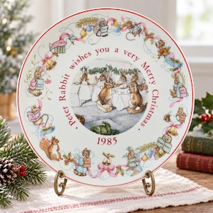 May include: A round white ceramic decorative plate featuring a central illustration of rabbits at a table. The border displays various small animals and holiday motifs with the text Peter Rabbit wishes you a very Merry Christmas 1985.