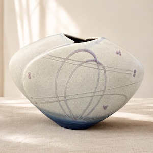 May include: A wide, rounded ceramic vase with a textured, off-white matte finish. The surface features etched, intersecting linear patterns in a soft purple hue, accented by small, delicate purple dot clusters. The base transitions into a deep blue.