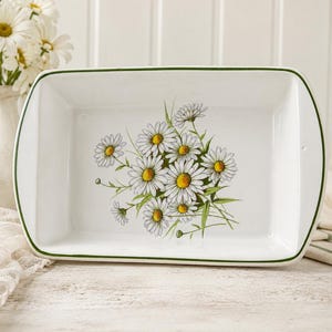 May include: A rectangular white ceramic baking dish featuring a central illustration of a cluster of white daisies with yellow centers and green stems. The dish has a thin green rim around the top edge and integrated handles on both sides.