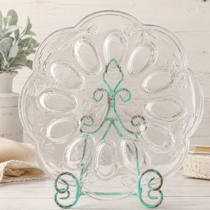Puede incluir: Una bandeja de cristal transparente para servir huevos con doce hendiduras ovaladas y borde festoneado, con detalles florales en relieve. La bandeja se apoya en un soporte de metal color verde azulado claro desgastado con volutas.