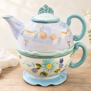 Puede incluir: Un juego de té individual de cerámica apilable que incluye tetera y taza. La tetera tiene un acabado en azul claro con flores naranjas pintadas y detalles dorados. La taza es de color crema con motivos florales azules y verdes.