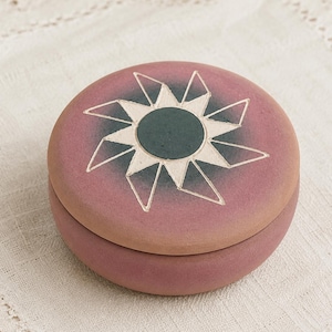 May include: A round, mauve-colored ceramic lidded box featuring a central dark green circle surrounded by a white, star-shaped geometric pattern on the lid. The container has a smooth, matte finish and a simple, cylindrical shape with a flat top.