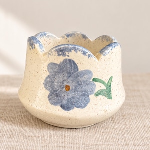 Peut inclure: Un pot en céramique avec une finition crème mouchetée et un bord festonné. L'extérieur présente une fleur bleue peinte à la main avec un centre jaune et une tige verte. Le bord est accentué par une texture bleue épongée, et la forme globale est arrondie avec une large ouverture.
