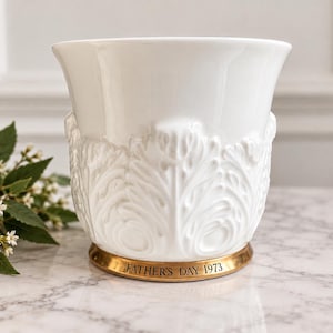 May include: A white ceramic vessel featuring a raised, embossed leaf pattern around the exterior. The base is encircled by a metallic gold-toned band with the black engraved text FATHER'S DAY 1973. The object has a wide, flared rim and a smooth finish.