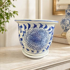 May include: A white ceramic planter with a blue hand-painted floral pattern featuring a large central blossom and leafy vines. The planter has a wide, flared rim and sits on a matching circular saucer, creating a cohesive two-piece set.