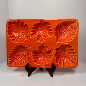 May include: A rectangular orange ceramic serving tray featuring six recessed compartments shaped like individual leaves. Each leaf section displays detailed embossed veining patterns across the surface, creating a textured appearance on the glossy, vibrant orange glazed finish.