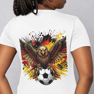 May include: A white short-sleeved t-shirt featuring a graphic on the back. The design displays a brown eagle with spread wings clutching a black and white football, set against a background of red, yellow, and black paint splatters.