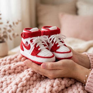 May include: A pair of handmade crochet baby booties designed to resemble high-top sneakers in a red and white color scheme. The booties feature white laces, a red ankle strap with a button, and a small circular logo with a basketball and the word BABY.