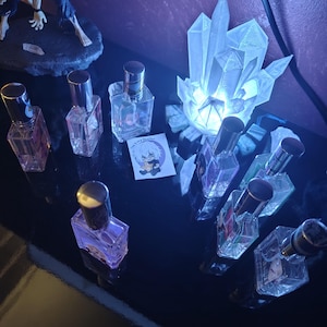 May include: Eight clear glass perfume bottles with silver caps arranged on a dark surface. A glowing white crystal lamp sits among the bottles, and a small square sticker featuring a cartoon character with white hair is placed in the center.