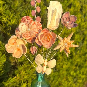 May include: A decorative bouquet of handcrafted flowers made from various seashells and pearls, featuring a central butterfly shell ornament, arranged in a small green glass bottle filled with light green sand, showcasing natural textures.