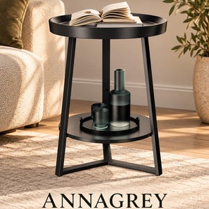 May include: A black metal side table with a circular top and a lower shelf. The table features three angled legs and a raised edge on the top surface. An open book rests on the top, while a tall glass vase and two small glass tumblers sit on the lower shelf.