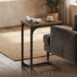 May include: A C-shaped side table with a dark brown wood-grain top and a black metal frame. The table holds an open magazine and a white ceramic coffee cup on a saucer. The text ANNAGREY INTERIORS is printed in gold at the bottom of the image.