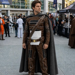 May include: A brown long-sleeved shirt and matching pants costume featuring a white rectangular chest panel, black accents, and a dark gray cape. A brown utility belt with a gold buckle and attached pouches is worn diagonally across the torso.