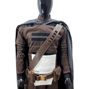 May include: A brown long-sleeved tunic with a white rectangular panel on the front, a dark gray cape, and a brown utility belt featuring a shoulder strap, a cylindrical pouch, and a rectangular flap pouch, all displayed on a black mannequin torso.