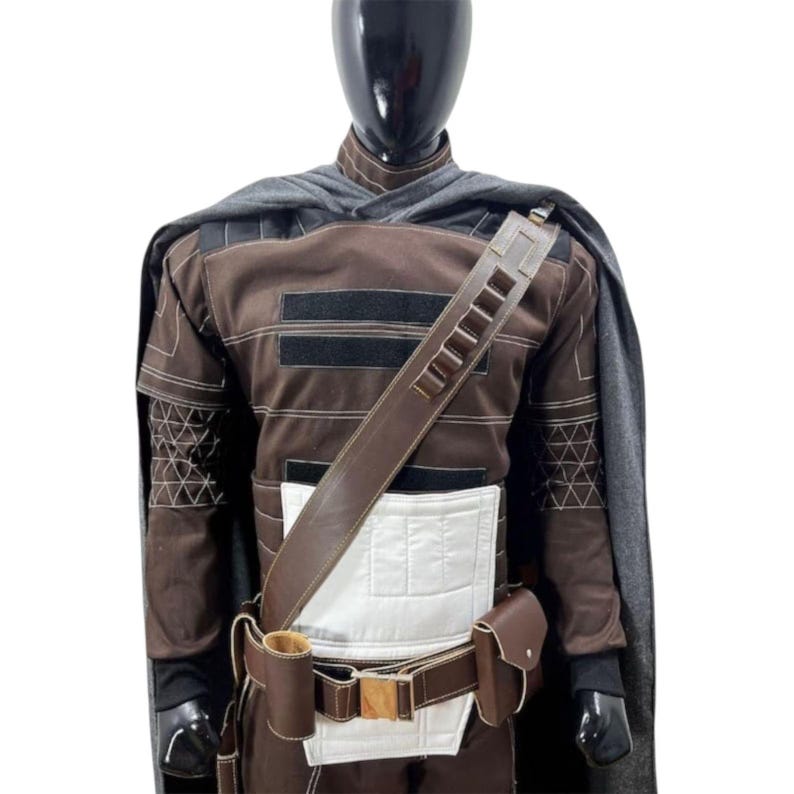 May include: A brown long-sleeved tunic with a white rectangular panel on the front, a dark gray cape, and a brown utility belt featuring a shoulder strap, a cylindrical pouch, and a rectangular flap pouch, all displayed on a black mannequin torso.