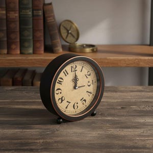 May include: A round analog table clock with a matte black frame and a copper-toned inner rim. The clock face is a light beige color with black Arabic numerals and ornate black metal hands. It stands on two small black spherical feet.