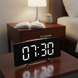 Può includere: Una sveglia digitale rettangolare con un quadrante nero curvo e numeri LED bianco brillante che indicano le 07:30. L'orologio presenta un design essenziale con una base in metallo argentato e pulsanti di controllo sul bordo superiore.