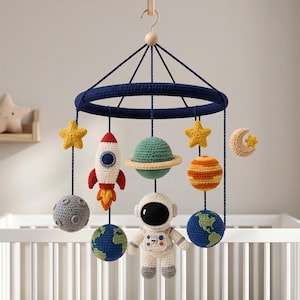 May include: A space-themed nursery mobile featuring a dark blue circular frame with hanging crocheted figures including an astronaut, a rocket ship, planets, stars, and a crescent moon. The figures are crafted in various colours like red, green, orange, and grey.