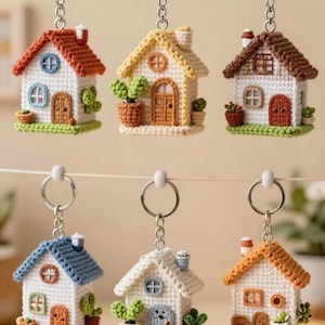 Peut inclure: Six porte-clés en forme de maison miniature réalisés en crochet, chacun avec un toit de couleur différente, une façade blanche, une petite cheminée et un anneau métallique. Les maisons sont ornées de petites plantes et portes.