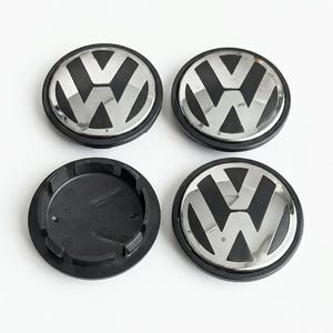 Könnte beinhalten: Ein Set aus vier runden Volkswagen Radnabendeckeln in Schwarz und Chrom mit dem VW-Logo. Drei Deckel sind von vorne abgebildet, während einer umgedreht ist, um die schwarze Kunststoffunterseite mit eingeprägtem Text zu zeigen.