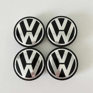 Könnte beinhalten: Vier runde Radnabendeckel mit dem Volkswagen-Logo in Schwarz und Silber. Jeder Deckel zeigt das ikonische VW-Emblem mit einem polierten silbernen V und W auf einem glänzend schwarzen Hintergrund, eingefasst von einem silbernen Rand.