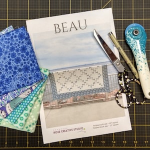 May include: A collection of quilting supplies including fabric, scissors, a rotary cutter, and a pattern titled "BEAU." The fabric swatches are various shades of blue and green. The scissors have a black and white polka dot pattern.