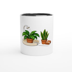 May include: A white ceramic mug with a black interior rim featuring a graphic illustration of a green leafy plant in a terracotta pot under a shower head, alongside a snake plant in a terracotta pot and a folded brown towel on a white surface.