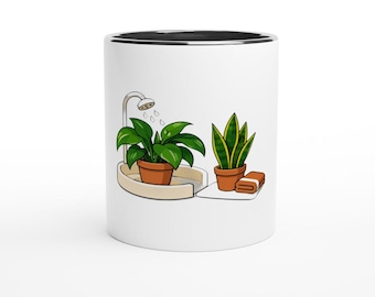 May include: A white ceramic mug with a black interior rim featuring a graphic illustration of a green leafy plant in a terracotta pot under a shower head, alongside a snake plant in a terracotta pot and a folded brown towel on a white surface.