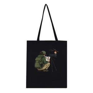 May include: A black tote bag featuring a graphic illustration of a person wearing a green cap, green hoodie, and tan pants, crouching while looking at a bird identification book next to a camera on a tripod with a small bird on a branch.