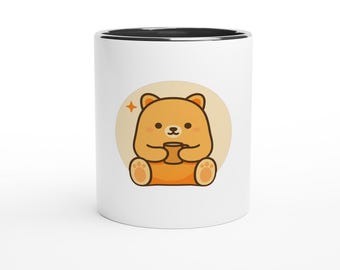 May include: A white ceramic mug with a black interior rim featuring a centered graphic of a light brown cartoon bear sitting down and holding a small brown mug. The bear is positioned inside a soft beige circle with a small orange star icon.