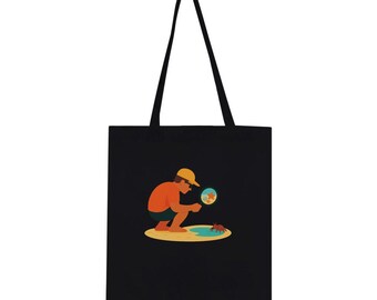 May include: A black tote bag featuring a graphic illustration of a person in an orange shirt and yellow cap crouching on a sandy beach. The person uses a magnifying glass to observe a small crab near a blue tide pool with starfish nearby.