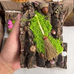 May include: A rectangular piece of dark brown tree bark decorated with bright green preserved moss, a small straw broom, a brown acorn, a miniature metal lantern, and several small brown seeds, all secured with thin pink thread.