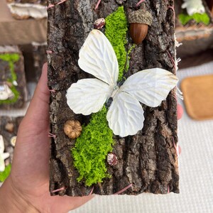 May include: A rectangular piece of dark brown tree bark decorated with a large white fabric butterfly, bright green preserved moss, a brown acorn with a cap, a small tan pumpkin-shaped bead, and two small patterned beans, stitched with pink thread.