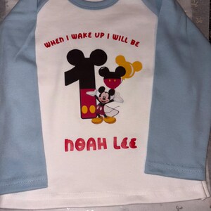 May include: A white long-sleeve shirt with light blue raglan sleeves. The front features a graphic of Mickey Mouse next to a large number one, a yellow balloon, and the text "WHEN I WAKE UP I WILL BE" and the name "NOAH LEE" in red lettering.