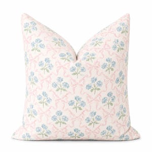 Può includere: Un cuscino quadrato con sfondo rosa chiaro caratterizzato da un motivo ripetitivo di piccoli bouquet di fiori blu e nastri rosa chiaro interconnessi. Il design è uniforme su tutta la superficie del rivestimento in tessuto.
