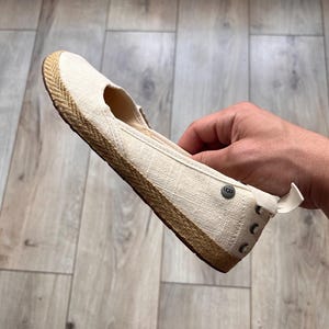 May include: A cream-colored canvas flat shoe with a textured jute-wrapped midsole. The shoe features a small round metal UGG logo stud on the side near the heel, a pull tab at the back, and a brown rubber outsole. The design is simple and low-profile.