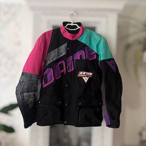 Puede incluir: Una chaqueta de moto negra con paneles de colores en rosa, turquesa y morado. La chaqueta muestra la palabra DAINESE en letras grandes de color morado en el pecho, un pequeño parche de DAINESE TECHNICAL WEAR y un patrón gris degradado.