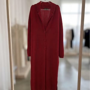 May include: A long-sleeved, knee-length red cardigan featuring a notched lapel collar, a button-down front with three visible buttons, and a straight hemline with side slits. The garment is crafted from a textured knit fabric and hangs on a hanger.