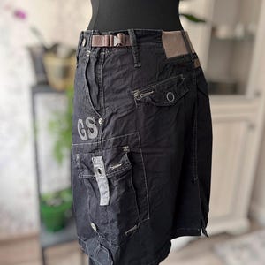 May include: A black cargo skirt featuring a brown adjustable waist strap, multiple utility pockets with button closures, and the letters GS embroidered in a light gray font on the front. The garment includes reinforced stitching and a side flap detail.