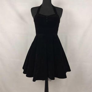 May include: A black velvet halter dress featuring a fitted bodice with three small buttons at the center neckline and a flared, knee-length skirt. The garment has a structured waistline and a sleeveless design, displayed on a mannequin.