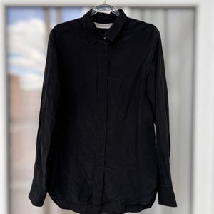 May include: A long-sleeved black button-down shirt featuring a classic collar, a concealed front placket, and buttoned cuffs. The shirt is displayed on a clear plastic hanger and includes a visible white interior neck label with black text.