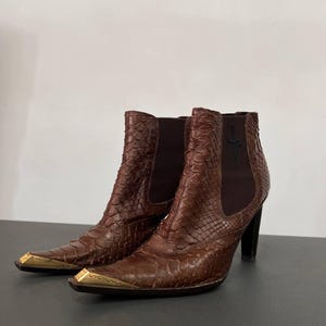 Puede incluir: Botines marrones con textura de piel de serpiente, con puntera afilada y punteras de metal dorado, paneles laterales elásticos de color marrón oscuro y tacón negro. Un pequeño emblema de cruz negra está bordado en el lateral de la caña.