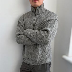 May include: A grey quarter-zip sweater featuring a chunky knit texture with subtle dark flecks throughout. The garment has a high ribbed collar, long sleeves with ribbed cuffs, and a ribbed hemline, providing a thick and warm appearance.