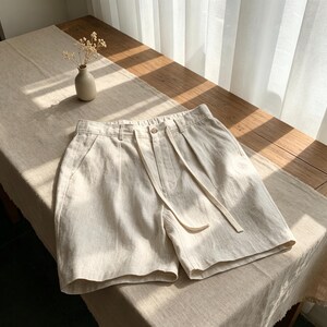 以下が含まれることがあります： ボタン留め、ジッパーフライ、共布のドローストリングタイを備えたハイウエストデザインのベージュのショートパンツ。両サイドにポケットがあり、フロントウエストには控えめなプリーツが施され、質感のある生地で作られています。