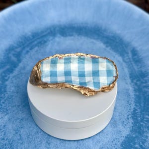 May include: A decorative oyster shell featuring a light blue and white gingham pattern on the interior surface. The edges of the shell are finished with a metallic gold trim, providing a contrast to the patterned center and the natural texture.